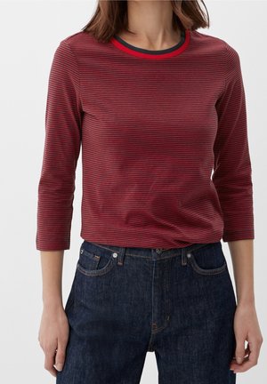 Frau trägt ein eng anliegendes rot-schwarz gestreiftes Langarmshirt mit Dreiviertelärmeln und dunkelblaue Jeans aus Denim.
