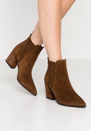 Braune Wildleder-Stiefeletten mit Blockabsätzen, getragen an nackten Beinen vor weißem Hintergrund.