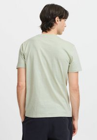 Tricou din bumbac de culoare verde deschis, cu guler rotund clasic, mâneci scurte și o textură simplă, văzut din spate. Design minimalist, fără modele vizibile.