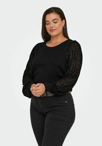 Crni pulover s rebrastim glavnim dijelom i prozirnim rukavima s leopard uzorkom. Uparen s crnim trapericama, koje imaju uski kroj i remen.