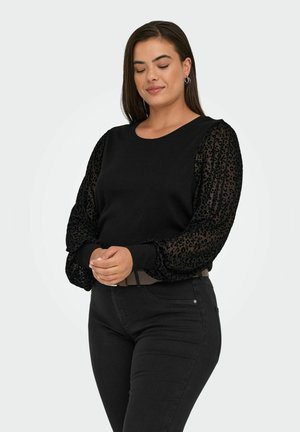 Schwarzer Pullover mit geripptem Hauptteil und durchsichtigen Puffärmeln im Leopardenmuster. Kombiniert mit schwarzen Jeans, die eine taillierte Passform und einen Gürtel haben.
