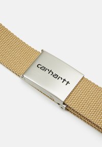 Curea din bumbac bej, cu cataramă metalică argintie, având logo-ul „carhartt” embosat. Curea texturată cu un finisaj neted și lustruit al cataramei.