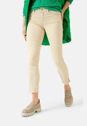 Jeans skinny beige con vita media, zip alle caviglie, abbinati a mocassini marroni chiari e una giacca trapuntata verde.