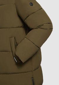 Olivovo zelená prešívaná bunda s kapucňou, ktorá obsahuje zips na bočnej vrecku a gumovou nášivku s logom na rukáve. Textúrovaná látka.