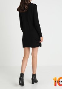 Abito nero a maniche lunghe con taglio dritto, caratterizzato da una sottile texture e dettagli con bottoni sulle spalle, abbinato a stivali neri alla caviglia.