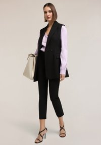 Vrouw met een zwarte mouwloze blazer, lila blouse, zwarte cropped broek, zwarte sandalen met bandjes, draagt een grote beige handtas, staand tegen een effen achtergrond.