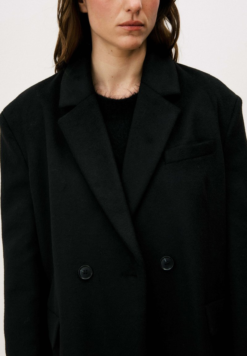 Manteau noir à double boutonnage en tissu texturé, avec col à revers, deux boutons et une poche poitrine. Aspect doux.