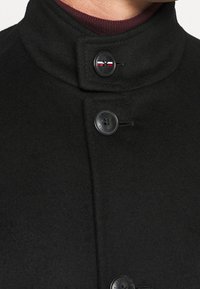 Cappotto di lana nera con colletto alto, dotato di due bottoni neri e un piccolo accento rosso, bianco e blu vicino al bottone superiore.