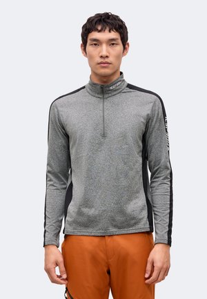 Un homme porte un haut de sport gris à fermeture éclair courte, avec des empiècements noirs et un pantalon orange, sur un fond clair et uni.