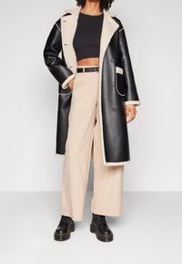 Manteau long en simili cuir noir avec doublure et finitions en shearling crème, coupe oversize, fermetures à boutons, associé à un pantalon large beige et des bottes.