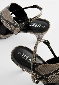 Sandales à talons hauts en imprimé serpent noir et beige avec un design de brides croisées et une semelle rembourrée. Présente un logo intérieur en texte blanc.