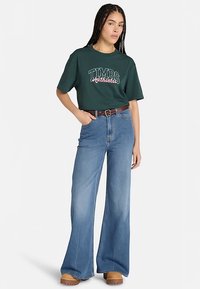 Ciemnozielona bawełniana koszulka z grafiką "TIMBS Authentic", w zestawieniu z jasnoniebieskimi szerokimi spodniami jeansowymi i beżowymi drewniakami na platformie, ozdobiona brązowym paskiem.