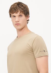 Tricou bej cu guler rotund, fabricat dintr-un material fin, având mâneci scurte și un mic logo negru brodat pe mâneca stângă.