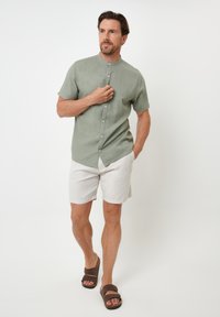 Kortærmet, knap-op skjorte i lysegrønt stof, parret med lyse beige shorts og brune sandaler, sat mod en hvid baggrund.