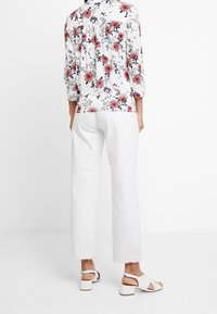 Bluse mit Blumenmuster in Weiß, mit Dreiviertelärmeln und gefälteltem Rücken, kombiniert mit weißen Caprihosen und hellen Sandalen.