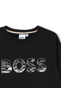 Črna trenirka z velikim belim napisom "BOSS" z valovito teksturo. Okrogla ovratnica z kontrastnim notranjim pasom.