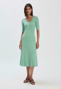 Rippstrickkleid in Hellgrün, mit V-Ausschnitt und kurzen Ärmeln. Der Saum ist leicht ausgestellt und hat eine strukturierte Oberfläche. Getragen mit braunen Sandalen.