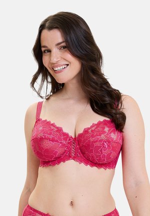 Donna sorridente con capelli castani lunghi che indossa un reggiseno in pizzo floreale rosa acceso, posando contro uno sfondo chiaro e semplice.