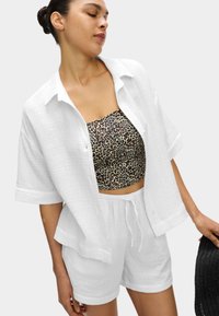 Camicia bianca a bottoni con texture sopra un crop top a stampa leopardata marrone e pantaloni bianchi. L'outfit presenta una vestibilità rilassata e un tessuto leggero.