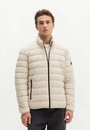 Junger Mann trägt eine hellbeige gesteppte Jacke mit schwarzem Reißverschluss und Taschen, steht vor einem schlichten weißen Hintergrund.