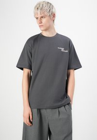 Junge Person mit kurzem blonden Haar, die ein dunkelgraues T-Shirt mit der Aufschrift "Trattoria Remo's" und eine lockere graue Hose trägt und zur Seite schaut.