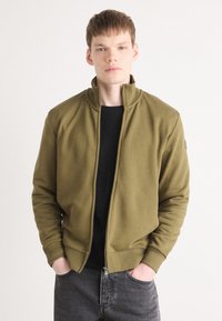 Olivinės žalios spalvos zip-up striukė su aukštu apykakle, gipiūrinėmis rankovėmis ir šoninėmis kišenėmis. Dėvima ant juodos megztinės, derinama su tamsiais džinsais.