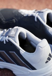 Par hvide og sorte atletiske løbesko med snørebånd på en struktureret overflade, der viser side- og hælmærkningsdetaljer.