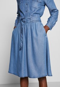 Jeansklänning i ljusblått, med långa ärmar, två bröstfickor, midja med bälte och utsvängd kjol med en fåll som går strax ovanför knäet.
