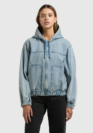 Junge Frau steht, trägt eine hellblaue Jeansjacke mit Reißverschluss und Kapuze mit Kordeln sowie schwarze Hose, vor einfachem Hintergrund.