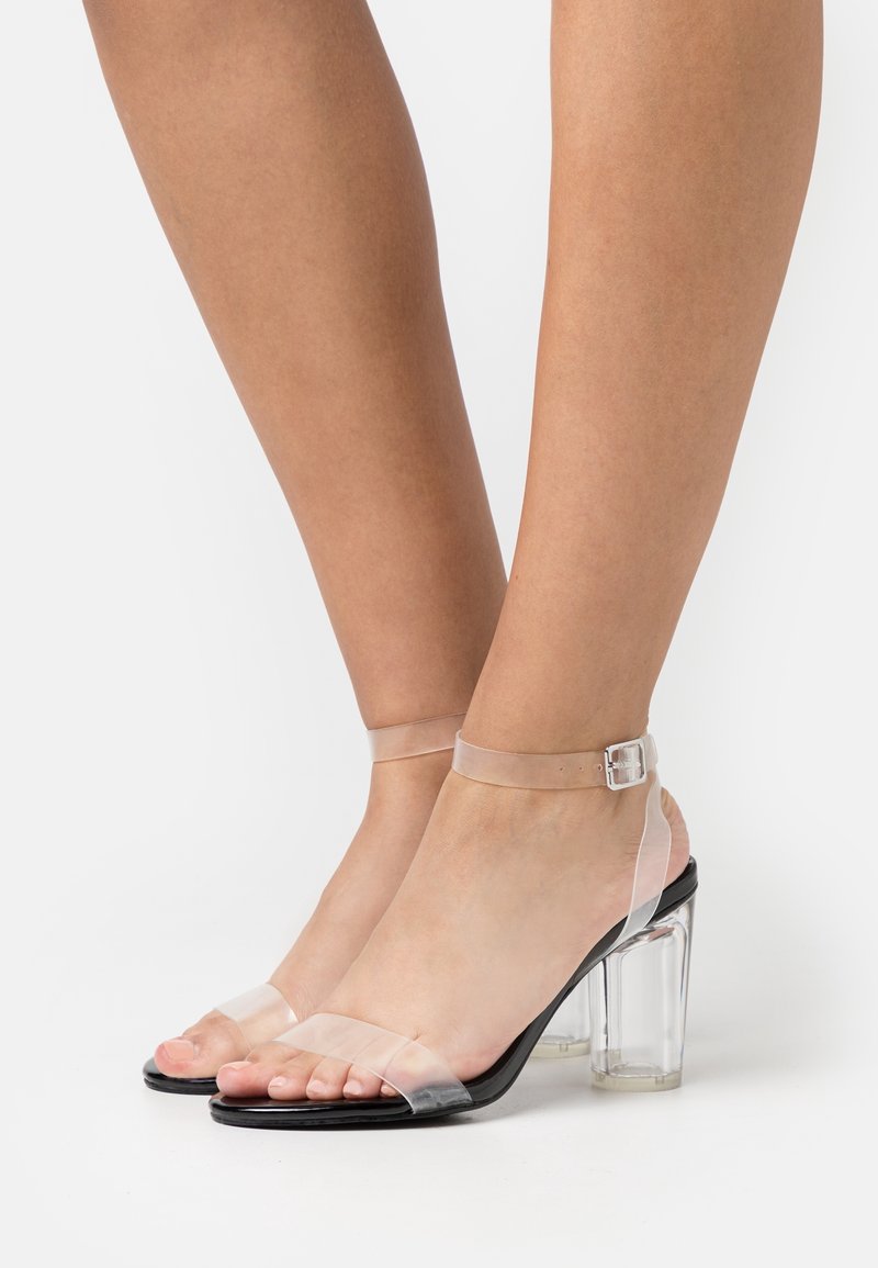 Pieds portant des sandales à talons hauts à bride cheville en PVC transparent avec semelles noires et talons blocs transparents sur fond blanc.