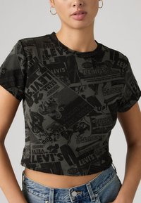 Μαύρο κοντό τοπ με υφασμένα σχέδια vintage διαφημίσεων. Στρογγυλή λαιμόκοψη και κοντά μανίκια. Συνδυασμένο με σορτς από τζιν.