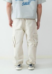 Beige cargobroek met meerdere zakken, losse pasvorm en een zachte textuur. Draag met grijze en witte sportschoenen. Eenvoudige achtergrond.