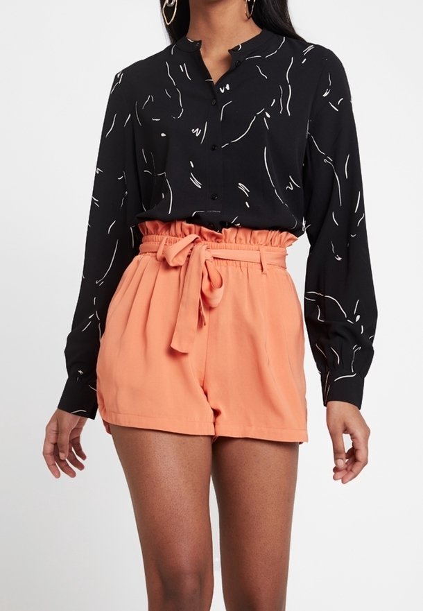 Femme portant une blouse noire à manches longues boutonnée avec des lignes abstraites blanches et un short pêche taille haute noué à la taille.