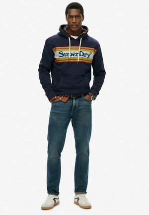 Sudadera con capucha azul marino con rayas blancas y naranjas, con un bolsillo tipo canguro, combinada con jeans azules y zapatillas blancas con suelas marrones.