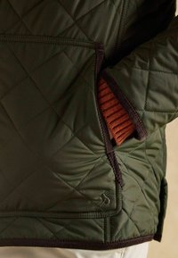 Gesteppte dunkelgrüne Jacke mit schwarzem Besatz und kleinem gesticktem Logo, eine Hand steckt in einer vorderen Tasche über einem rostfarbenen Ärmel.