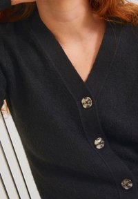 Cardigan nero in tessuto testurizzato, con scollatura a V e tre bottoni tondi decorati lungo la parte anteriore.