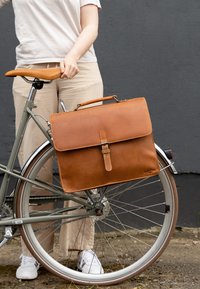 Bruine leren schoudertas met een klep en een gesp-sluiting, bevestigd aan een lichtgroene fiets. Beschikt over een gladde textuur en minimale stiksels.