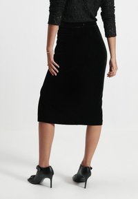 Jupe midi en velours noir avec une silhouette ajustée, dotée d'une fente au dos et portée avec des chaussures à talons noires. Haut noir texturé, à manches longues.