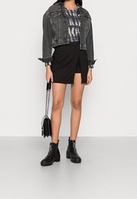 Chaqueta de mezclilla negra corta, camiseta de estampado tie-dye, falda envolvente negra y botas negras de tobillo. Pequeño bolso con detalles metálicos. Texturas suaves.