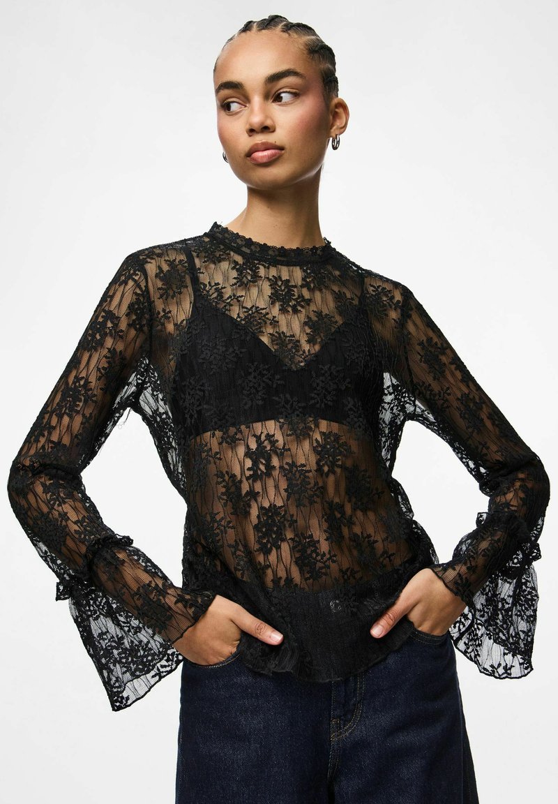 Crni top dugih rukava od čipke s cvjetnim uzorcima, prozirnom teksturom, naboranim manšetama i okruglim izrezom. Nošen preko crnog kamizola.