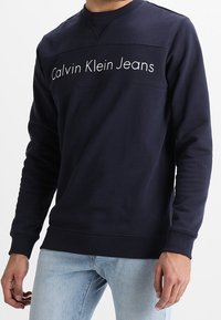 Felpa blu navy con scollo tondo, caratterizzata dalla scritta "Calvin Klein Jeans" bianca sul petto. Realizzata in morbido tessuto misto cotone.