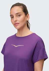 Lila Sport-T-Shirt mit kurzen Ärmeln, rundem Halsausschnitt und einem goldenen Logo-Akzent auf der Brust. Hergestellt aus einem glatten, atmungsaktiven Material.
