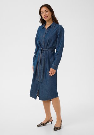 Femme portant une robe-chemise en jean à manches longues et ceinturée, avec des escarpins à imprimé zèbre, debout sur un fond blanc uni.