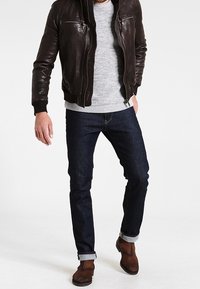 Giacca bomber in pelle marrone sopra un maglione grigio a maglia, jeans blu scuro con risvolti arrotolati e stivaletti marroni. Vestibilità casual e design moderno.