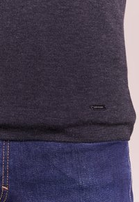 Mörkgrå stickad tröja med en texturerad yta, försedda med en svart märkesskylt. Bärs över blå denimjeans med synlig sömnad.