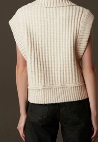 Pull sans manches beige en tricot avec un col montant, présentant une texture côtelée verticale et un ourlet ajusté. Un jean foncé est visible en dessous.