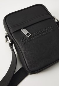 Černá crossbody taška vyrobená z texturované kůže, s zipem a raženým logem na přední straně. Popruh je plochý a nastavitelný.