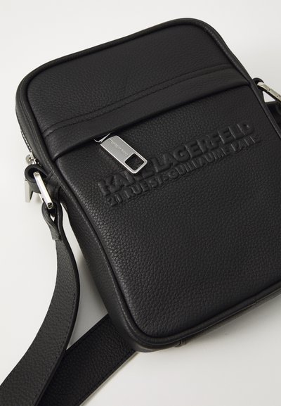 Sac bandoulière noir en cuir texturé, avec une fermeture éclair et un logo en relief sur le devant. La sangle est plate et réglable.