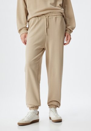 Beige Joggerhosen aus einem weichen Stoff mit einem elastischen Bund und Bündchen. Kombiniert mit weißen Sneakers, die ein beige-weißes Farbschema aufweisen.