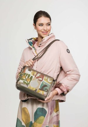 Woman in a pink puffer jacket smiling, holding a colorful patterned handbag with nature-inspired designs against a plain light background.
